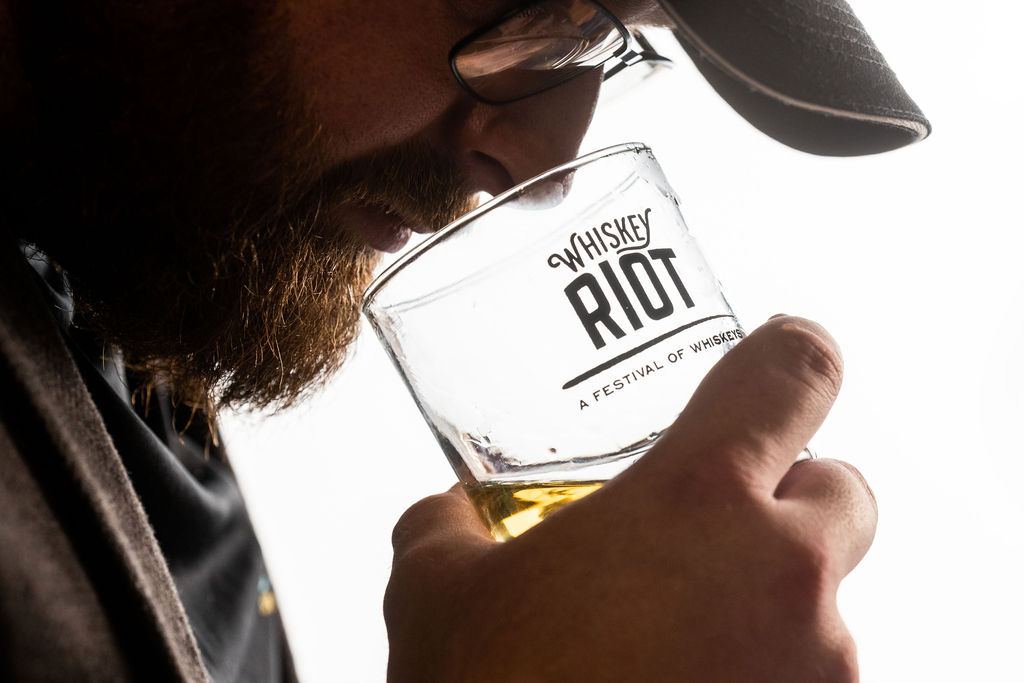 A close-up of a person smelling a whiskey drink in a clear glass branded with 'Whiskey Riot: A Festival of Whiskeys'.