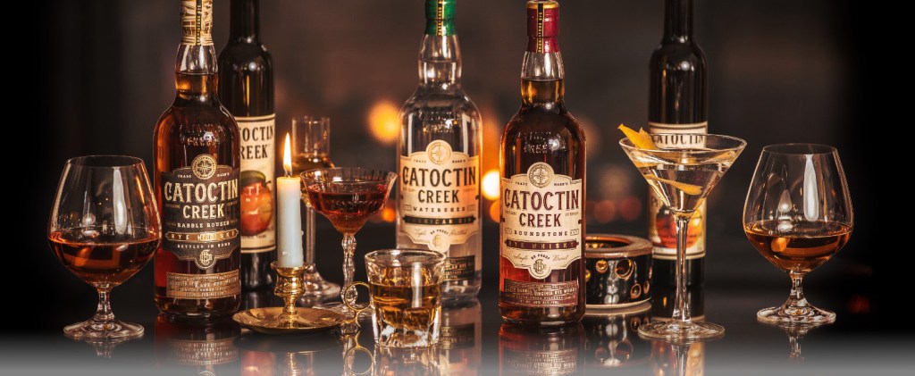 A collection of Catoctin Creek spirits displayed on a reflective surface, including various bottles and glasses filled with different types of drinks, in front of a softly lit background.