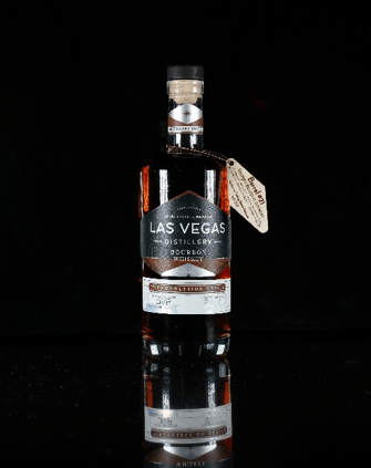 A bottle of Las Vegas distillery whiskey with a black label and a tag, placed on a reflective surface against a black background.