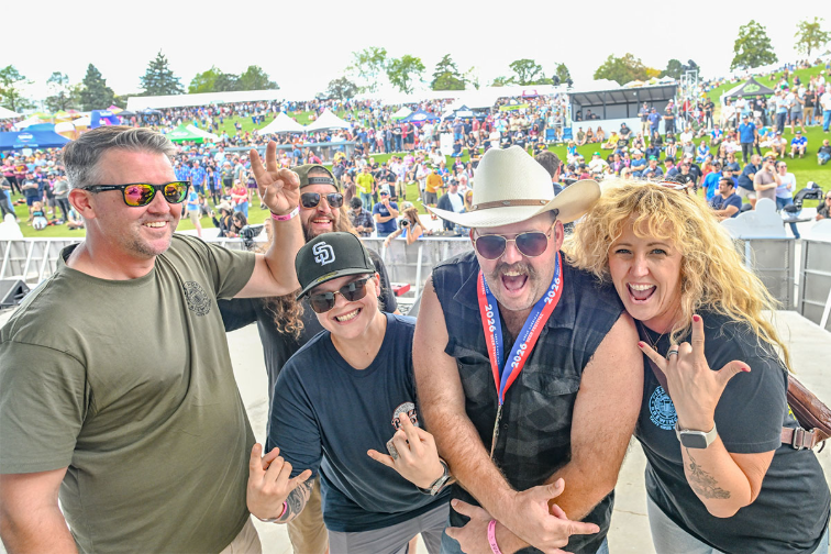 A group of five people posing joyfully in front of a large outdoor crowd at a festival, with vibrant tents and greenery in the background.